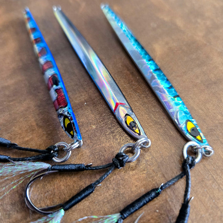 Close-up of three Submission Fishing Ogre Jr 10g slow pitch jigs in different color variations with assist hooks displayed on a wooden surface.
