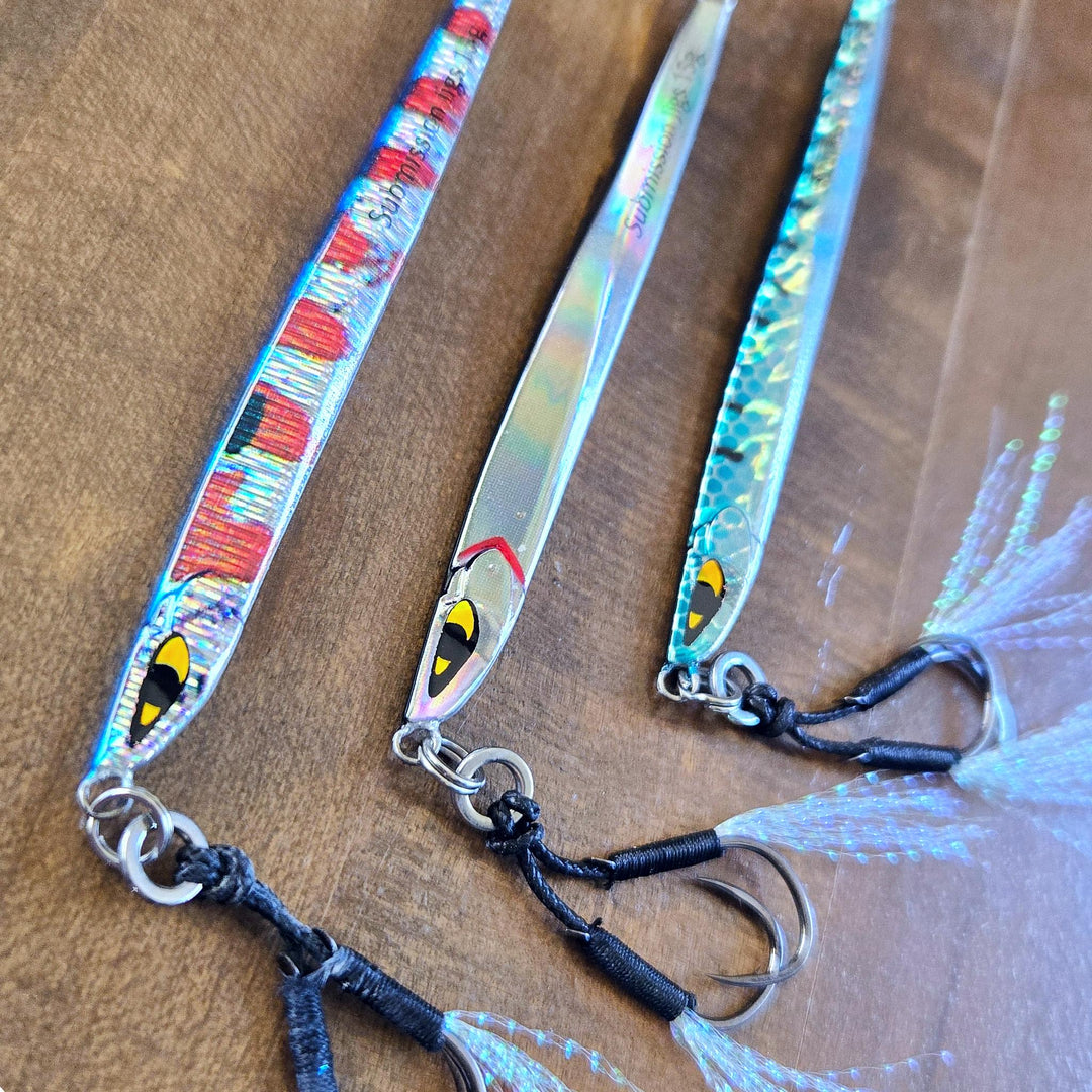 Close-up of three Submission Fishing Ogre Jr 15g slow pitch jigs in different color variations with assist hooks displayed on a wooden surface.